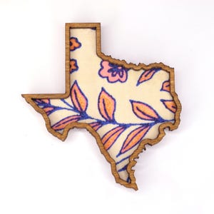 May include: Wooden Texas-shaped decorative item with a floral design. The interior features a cream-colored background with pink flowers and orange and blue leaves. The border is a natural wood color.
