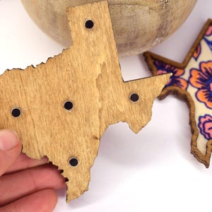 May include: Wooden Texas-shaped magnets with black circular magnets. One magnet is plain wood, and the other features a floral design with orange, pink, and blue colors. The magnets are likely for a refrigerator or a magnetic board.