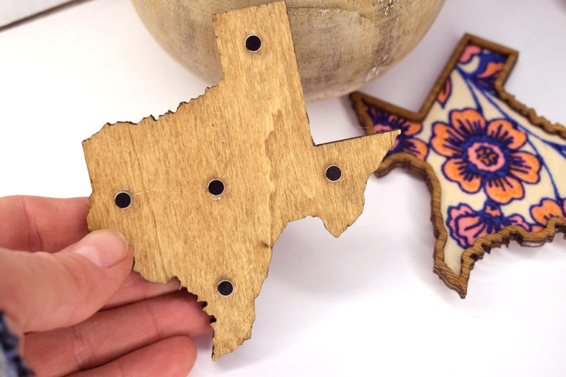 May include: Wooden Texas-shaped magnets with black circular magnets. One magnet is plain wood, and the other features a floral design with orange, pink, and blue colors. The magnets are likely for a refrigerator or a magnetic board.