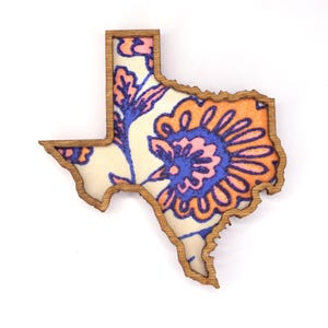 May include: Wooden Texas-shaped decorative item with a floral pattern. The design features a cream-colored background with orange, blue, and pink floral elements. The outline of the state is a natural wood color.
