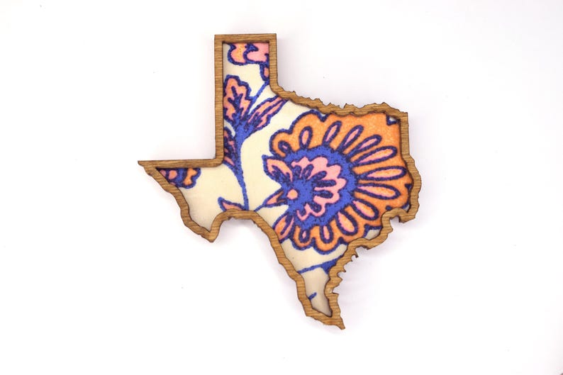 May include: Wooden Texas-shaped decorative item with a floral pattern. The design features a cream-colored background with orange, blue, and pink floral elements. The outline of the state is a natural wood color.