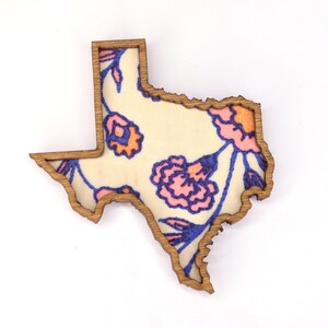 May include: Wooden Texas-shaped decorative piece with a floral design. The interior features a cream-colored background with pink, orange, and blue floral patterns. The outline is a natural wood color.
