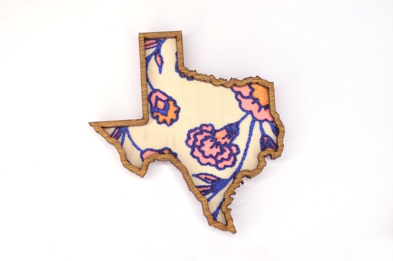 May include: Wooden Texas-shaped decorative piece with a floral design. The interior features a cream-colored background with pink, orange, and blue floral patterns. The outline is a natural wood color.