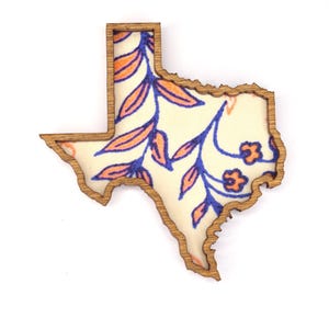 May include: Wooden Texas-shaped cutout with a floral design. The cutout has a natural wood border and features a cream-colored interior with blue and orange floral patterns. The design includes leaves and small flowers.