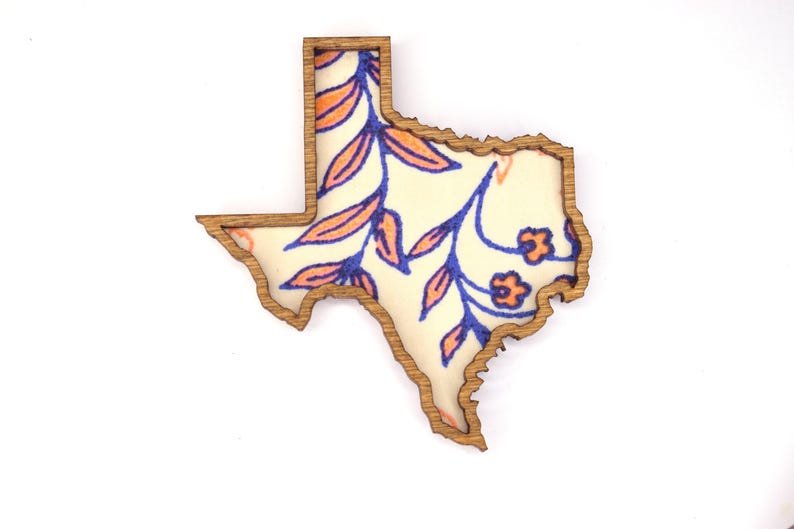 May include: Wooden Texas-shaped cutout with a floral design. The cutout has a natural wood border and features a cream-colored interior with blue and orange floral patterns. The design includes leaves and small flowers.