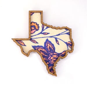 May include: Wooden Texas-shaped decorative item with a floral design. The outline is a light brown, and the interior features a cream-colored background with blue and orange floral patterns. The item is likely a decorative piece or a souvenir.