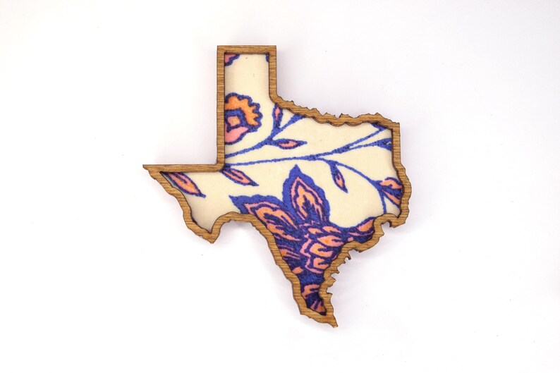 May include: Wooden Texas-shaped decorative item with a floral design. The outline is a light brown, and the interior features a cream-colored background with blue and orange floral patterns. The item is likely a decorative piece or a souvenir.