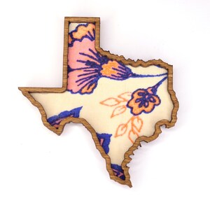 May include: Wooden Texas-shaped decorative item with a floral design. The interior features a cream-colored background with pink, blue, and orange flowers. The wooden frame has a natural wood grain finish.