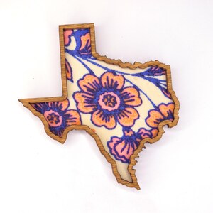 May include: Wooden Texas-shaped decoration with a floral design. The flowers are orange and pink with blue accents, set against a cream background. The border is a natural wood color.