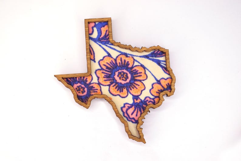 May include: Wooden Texas-shaped decoration with a floral design. The flowers are orange and pink with blue accents, set against a cream background. The border is a natural wood color.
