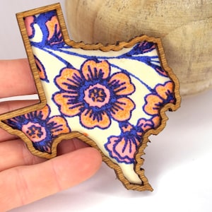May include: Wooden Texas-shaped decorative item with a floral design. The design features orange, blue, and pink flowers on a cream background. The wooden border is a light brown color.