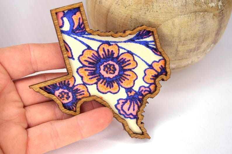 May include: Wooden Texas-shaped decorative item with a floral design. The design features orange, blue, and pink flowers on a cream background. The wooden border is a light brown color.