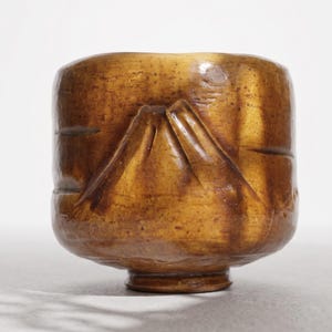 May include: A cylindrical ceramic tea bowl with a textured, mottled golden-brown glaze. The exterior surface features a prominent, raised triangular relief design resembling a mountain peak, complemented by horizontal carved grooves.