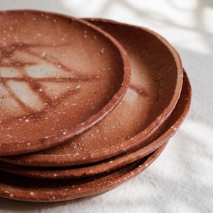 May include: A stack of four handmade ceramic plates. The plates are a reddish-brown color with a speckled texture and a slightly raised rim. Each plate has a unique, light-colored pattern in the center.