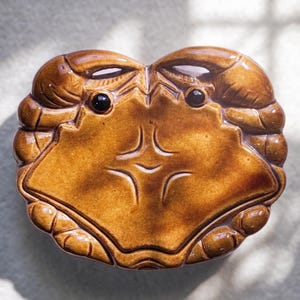 May include: A ceramic crab-shaped dish with a glossy, brown finish. The dish features detailed claw and body segments, with two black, round eyes. The crab's face has a stylized design. The dish is likely used for serving or decoration.