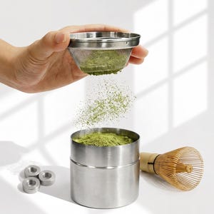 Op de afbeelding: Een roestvrijstalen matcha-zeef wordt boven een cilindrische roestvrijstalen container gehouden, waarbij groen matchapoeder in de container valt. Een bamboe garde en drie kleine metalen ringen liggen naast de container op een wit oppervlak.