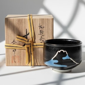 Könnte beinhalten: Eine schwarze Keramik-Teetasse mit einem Bergdesign in Weiß und Blau, neben einer Holzkiste, die mit einem gelb-schwarzen Band gebunden ist. Die Box hat japanische Schriftzeichen.