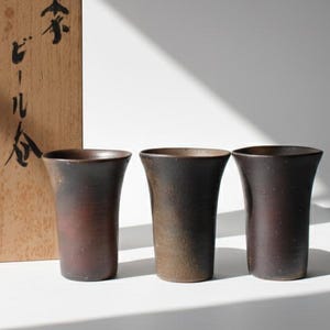 Puede incluir: Tres tazas de cerámica marrón oscuro con la parte superior acampanada se exhiben junto a una caja de madera con texto japonés. Las tazas tienen un aspecto rústico y texturizado. La caja de madera tiene caracteres japoneses y un sello rojo.
