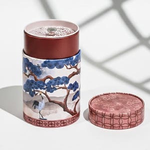May include: A cylindrical container with a lid, featuring a watercolour design of a tree, a crane, and a landscape in blue, brown, and white. The lid and top section are a deep red colour. A separate, smaller, round container with a patterned lid is also present.