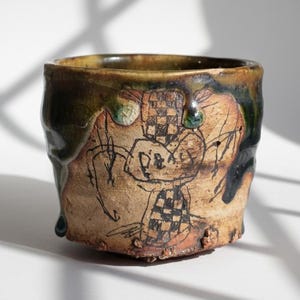 May include: Handmade ceramic cup with a unique, textured surface. The cup features a dark green and brown glaze around the rim, with black and brown drawings on the beige body. The design includes a checkered pattern and the word "Peace".
