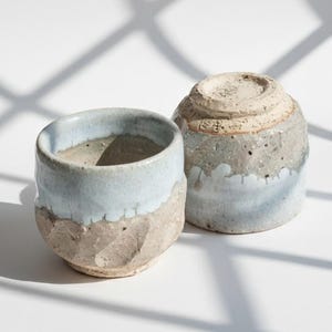 Könnte beinhalten: Zwei handgefertigte Keramikbecher. Ein Becher steht aufrecht und zeigt einen hellblauen Rand und eine strukturierte, hellbraune untere Hälfte. Der andere Becher steht auf dem Kopf und zeigt ein ähnliches Farbschema. Die Becher stehen auf einer weißen Oberfläche mit Schatten.
