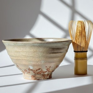 Puede incluir: Un cuenco de matcha de cerámica con un esmalte marrón claro y beige, y un batidor de matcha de bambú. El cuenco tiene una forma ancha y poco profunda con una superficie texturizada. El batidor tiene un mango dorado.
