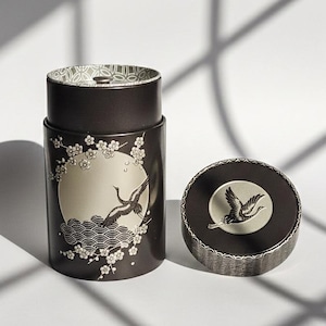 May include: A dark brown cylindrical container with a lid, featuring a crane design. The container has a silver-colored circle with a crane illustration, surrounded by cherry blossoms and waves. The lid also has a crane design.