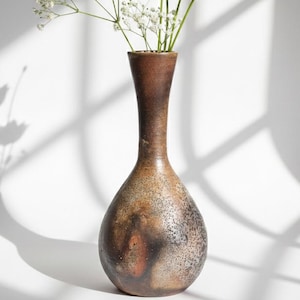 May include: A brown ceramic vase with a slender neck and a rounded base. The vase has a speckled, textured finish with dark brown and reddish-brown tones. White flowers and foliage are arranged inside the vase, with shadows on the white backdrop.