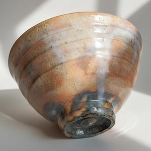 Pode incluir: Uma tigela de cerâmica artesanal com uma superfície texturizada única. A tigela apresenta uma mistura de tons terrosos, incluindo castanho, laranja e branco, com uma base escura. O design da tigela mostra uma estética rústica.