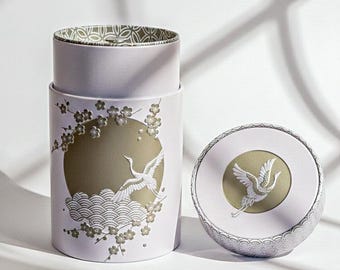 May include: A lavender cylindrical container and lid, decorated with a gold circle and a crane design. The container has a detailed relief of a crane, waves, and cherry blossoms. The lid also features a crane design.