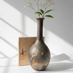 May include: A tall, dark brown ceramic vase with a bulbous base and slender neck, holding delicate white flowers. The vase has a textured, metallic finish with gold accents. A wooden box with Japanese calligraphy is behind the vase.