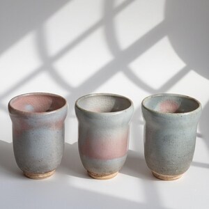 May include: Three handmade ceramic cups. Each cup has a unique color gradient, with a light blue base and a pink rim. The cups have a textured surface and a simple, elegant design, perfect for serving beverages.