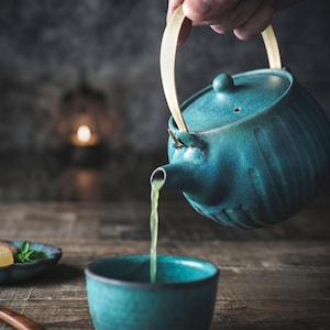 May include: A teal ceramic teapot with a ribbed texture and a light-colored bamboo handle pouring green tea into a matching teal ceramic cup. The teapot features a lid with a small knob and a curved spout, showcasing a consistent glaze finish.