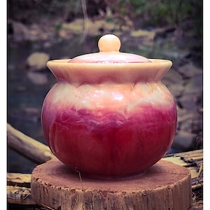 May include: A small, round, lidded jar with a gradient of red and white hues. The jar sits on a wooden slice, with a natural background of a stream and foliage. The lid has a small, round knob. The jar is likely for decorative or storage purposes.