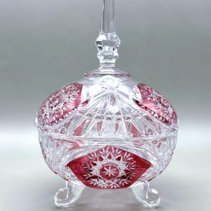 Pode incluir: Uma bomboniere redonda de cristal transparente com tampa e três pés pequenos. O prato apresenta um design de vidro lapidado vermelho rubi e transparente com um padrão de estrela. A tampa tem um pináculo alto e pontiagudo. Uma peça decorativa para a casa.