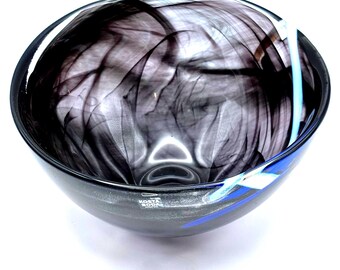 May include: A round glass bowl with a dark gray and black swirled design. The bowl has a wide opening and a dark rim with blue and white accents. The brand name "KOSTA BODA" is visible on the side.