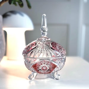 Może przedstawiać: Przezroczysty, szlifowany szklany pojemnik na cukierki z rubinowoczerwonym wnętrzem i akcentami. Naczynie ma okrągły korpus, pokrywę z wysokim, spiczastym zwieńczeniem i trzy małe nóżki. Szlifowane szkło ma geometryczne wzory i detale w kształcie gwiazd.