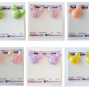 Op de afbeelding: Zes paar kleurrijke glitter Mickey Mouse-vormige oorbellen. De oorbellen worden getoond op witte kaartjes met de tekst "Enchantment Jewelry" en social media-handvatten.