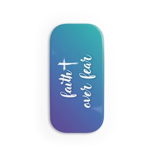 Puede incluir: Un imán rectangular con esquinas redondeadas, con un degradado de azul y morado. La escritura blanca dice "faith + over fear" con un símbolo de cruz. El imán es un artículo religioso o inspirador.