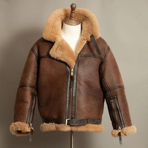 Puede incluir: Chaqueta de cuero marrón con cuello y forro de borrego beige. La chaqueta tiene cierre de cremallera, cinturón en la cintura y bolsillos con cremallera en las mangas. La chaqueta se exhibe en un maniquí.