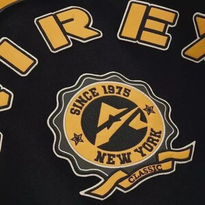 K&ouml;nnte beinhalten: Schwarzer Stoff mit gelben und wei&szlig;en gestickten Aufn&auml;hern. Die Aufn&auml;her zeigen das Wort "REX" und ein rundes Emblem mit "SINCE 1975 NEW YORK CLASSIC" und einem Blitz.
