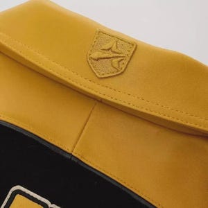 K&ouml;nnte beinhalten: Nahaufnahme eines senfgelben Lederartikels mit einem aufgestickten Emblem. Das Emblem hat die Form eines Schildes mit einem aufgestickten Design. Der Artikel hat einen schwarzen Abschnitt mit einem wei&szlig;en und gelben Design.