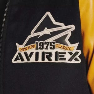 K&ouml;nnte beinhalten: Nahaufnahme einer schwarz-gelben Jacke mit einem Aufn&auml;her. Der Aufn&auml;her zeigt den Text "NEW YORK 1975 CLASSIC AVIREX" in Wei&szlig;, Gelb und Schwarz. Die Jacke hat einen schwarzen K&ouml;rper und gelbe &Auml;rmel.
