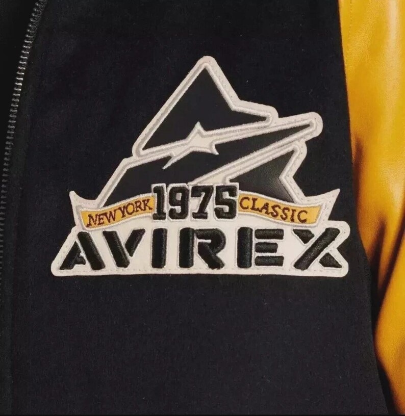 K&ouml;nnte beinhalten: Nahaufnahme einer schwarz-gelben Jacke mit einem Aufn&auml;her. Der Aufn&auml;her zeigt den Text "NEW YORK 1975 CLASSIC AVIREX" in Wei&szlig;, Gelb und Schwarz. Die Jacke hat einen schwarzen K&ouml;rper und gelbe &Auml;rmel.