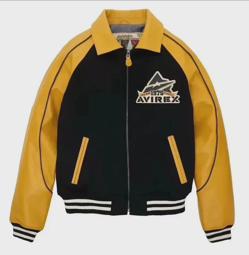 K&ouml;nnte beinhalten: Schwarze und gelbe Bomberjacke mit dem Text "AVIREX 1975 CLASSIC" auf der Brust. Die Jacke hat einen Rei&szlig;verschluss, einen Kragen und gestreifte B&uuml;ndchen und Saum. Die &Auml;rmel sind gelb und der K&ouml;rper ist schwarz.