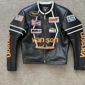 Puede incluir: Chaqueta de moto de cuero negro con múltiples parches, incluyendo una bandera americana, "Champion" y "X-Ray". La chaqueta presenta rayas blancas en las mangas y el nombre "Vanson" en letras naranjas. Una chaqueta clásica de estilo racing.
