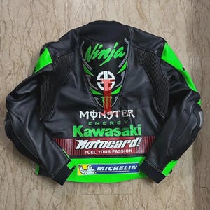 May include: Black and green motorcycle jacket with "Ninja" and "Monster Energy Kawasaki" logos. The jacket features a "Motocard" logo and "Fuel Your Passion" text, along with a Michelin logo. The jacket has green accents on the shoulders and sleeves.
