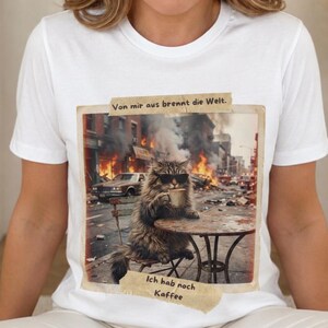May include: A white t-shirt featuring a graphic print of a fluffy cat wearing sunglasses, sitting at a table with a coffee mug amidst a chaotic city street scene with fire and smoke. The text reads: Von mir aus brennt die Welt. Ich hab noch Kaffee.