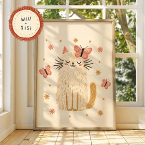 May include: Embroidered artwork of a cat with a butterfly on its head and two butterflies nearby. The cat is beige with black whiskers and a smiling face. The butterflies and flowers are pink and gold. The artwork is framed and has the text "Will & Sisi".