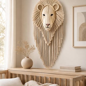 May include: A wall-mounted lion head sculpture crafted from cream-colored macrame cord. The design features a detailed face with dark eyes and a pink nose, surrounded by a thick, layered fringe mane that extends downward in a long, pointed shape.
